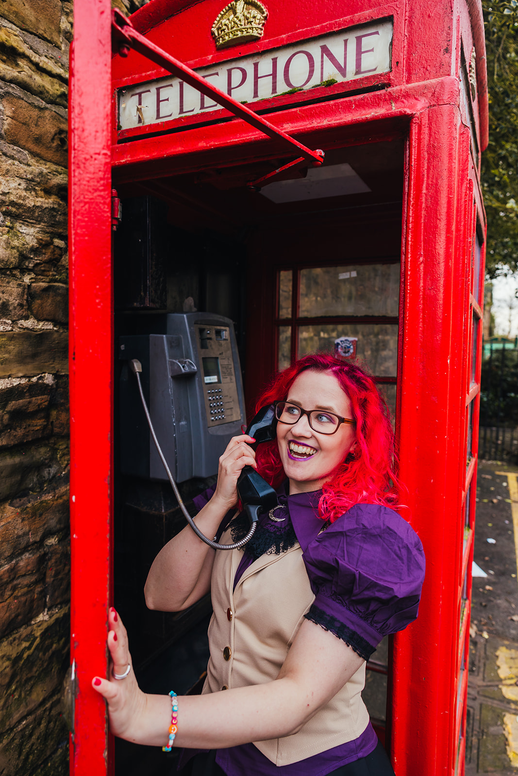 Natalie stands in the doorway of a traditional red telephone box, holding the black phone to her ear and with a big smile on her face.